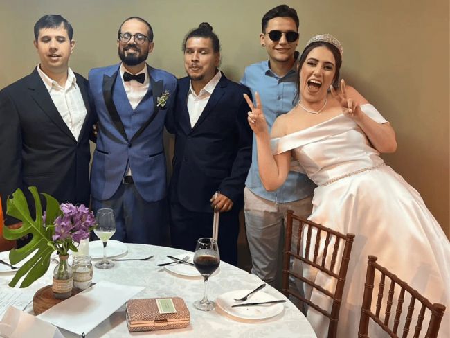 Da esquerda para direita: Daniel está usando um terno preto e blusa rosa claro, Pulis vestindo um terno azul jeans, Renan um terno preto e blusa branca com um coque de samurai, Giovanni está de blusa social azul clara e calça beje e óculos escuros, Mari está celebrando fazendo uma careta de felicidade. Na mesa está o cardápio em braile.