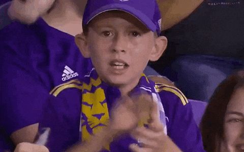 Garoto comemorando na arquibancada de um time de futebol. Ele está vestido com uma blusa azul e boné. Gesticula com as mãos vibrando e dizendo Sim, vamos lá
