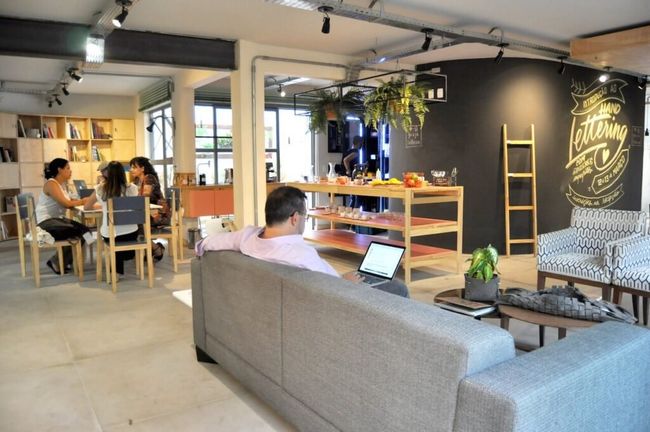 Entrada do Guaja Coworking. Na imagem existem três mulheres sentadas ao fundo em uma mesa de madeira clara e um homem com um notebook aberto em um sofá.