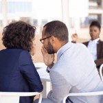 Um homem sussurando ao ouvido de uma mulher comentando sobre um colega de trabalho